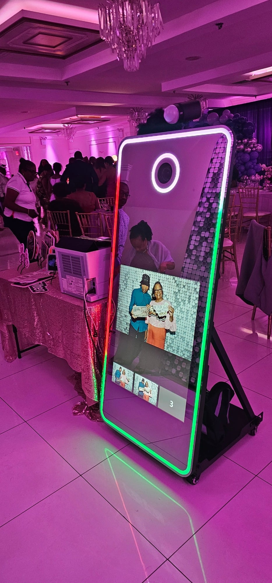 A vertical photo booth mirror with colorful LED lights displays a photo of two people in a banquet hall filled with guests.