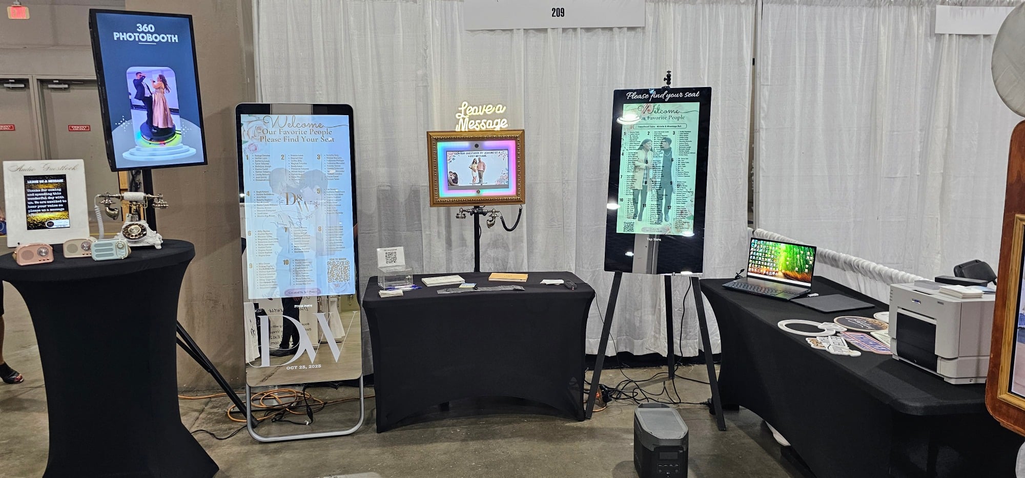 Event display featuring a 360 photo booth, interactive mirror, framed photo station, and laptop with printer on black tables.