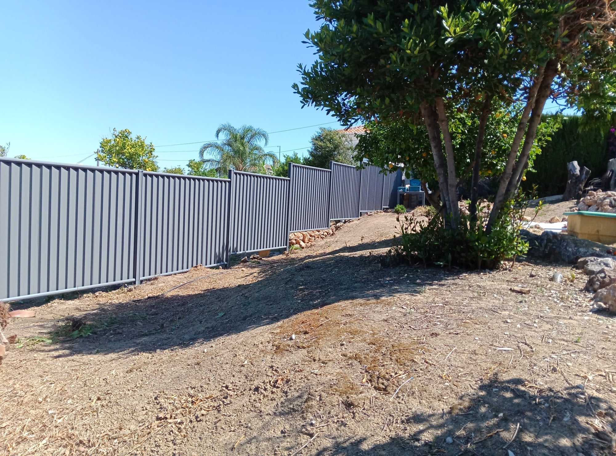 Grey ColourFence steel fence with vertical panels and matching posts along a garden boundary beside mature trees.